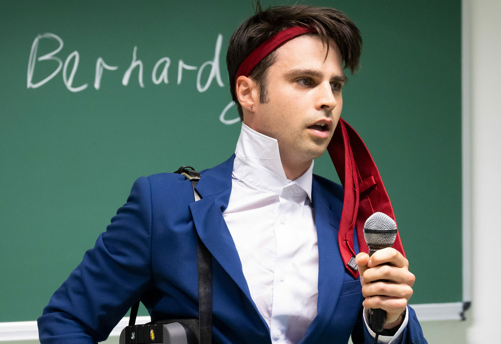 Eric Bernardy hat sich seinen Schlips über die Strin gebunden und spricht in das Handmikrofon seines Kassettenrekorder, den er in einer Tragetasche über der Schulter trägt.