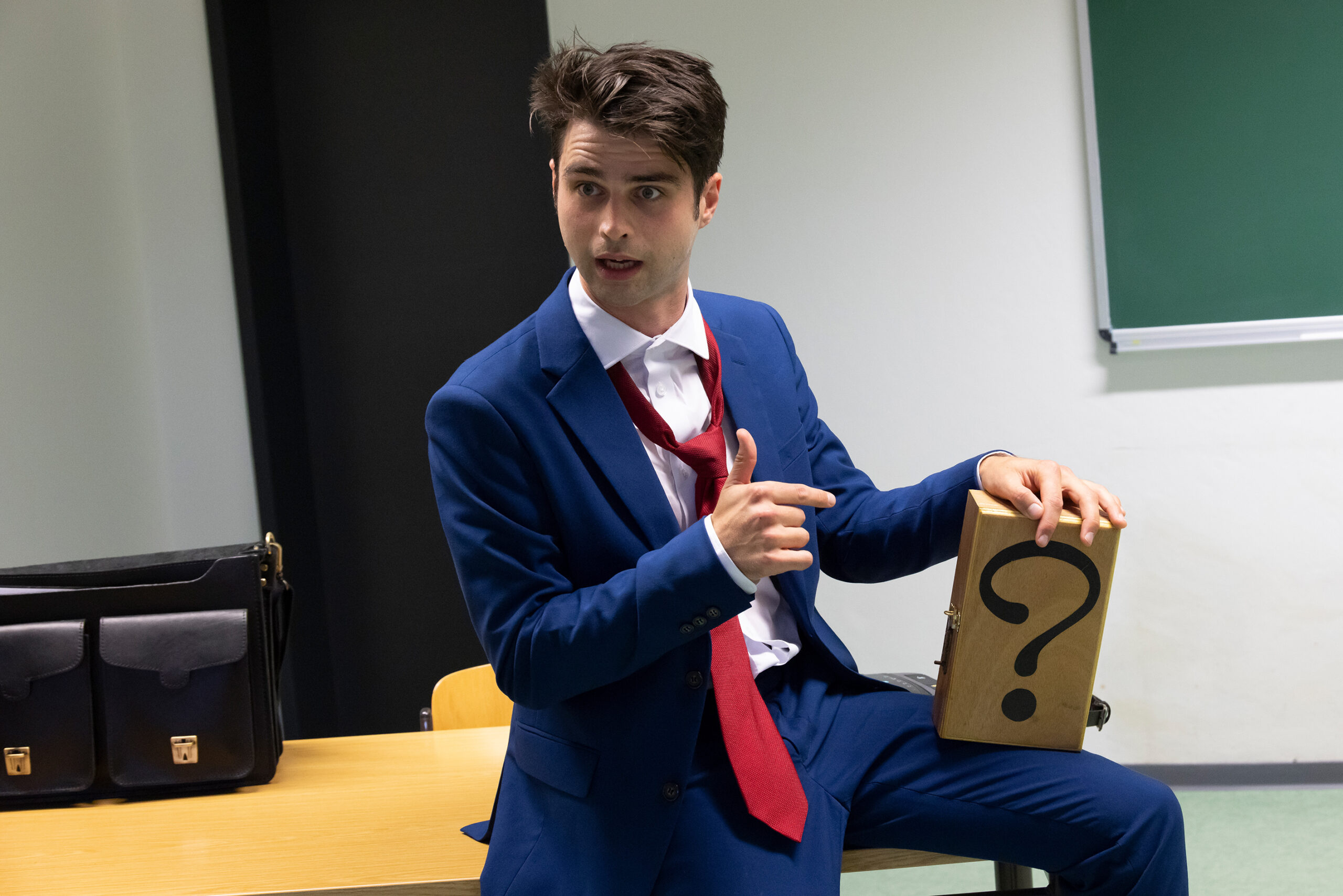 Eric Bernardy sitzt jovial auf einem Lehrertisch und hält eine Holzbox mit einem Fragezeichen drauf auf seinem Oberschenkel. Er zeigt mit erklärender Geste auf die Box.