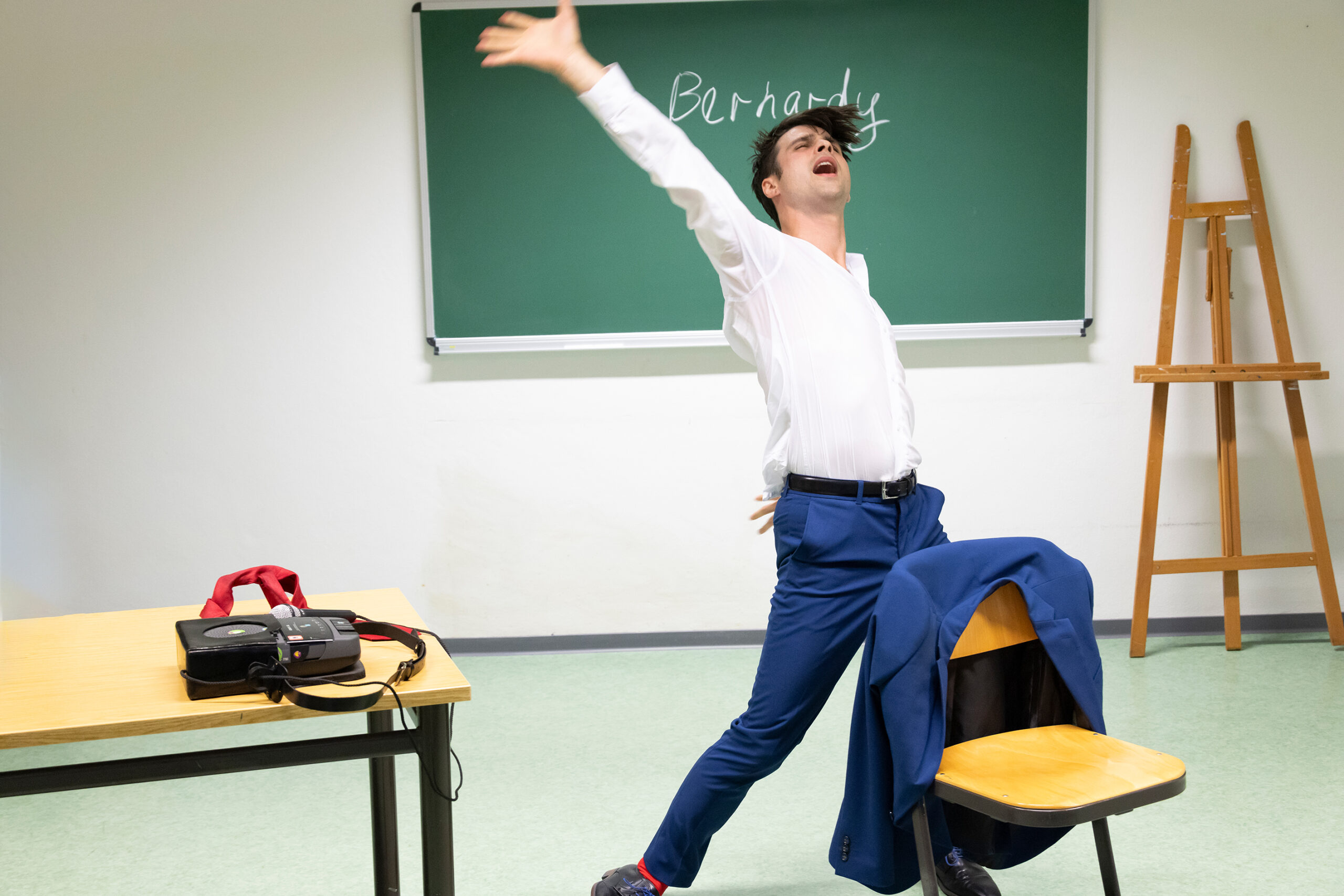 Eric Bernaredy wirft vor einer grünen Tafel in einem Klassenzimmer seine Arme in die Höhe als würde er expressiv tanzen.